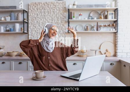 Une jeune femme musulmane freelance, étudiante, femme d'affaires dans un hijab est assise à la maison dans la cuisine en écoutant de la musique dans les écouteurs d'un ordinateur portable. Elle se repose, se détend, danse, lève les mains. Banque D'Images