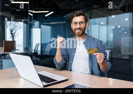 Un jeune homme utilise avec succès une carte de crédit. Il s'assoit heureux dans le bureau à l'ordinateur, tient une carte, regarde la caméra, montre un geste oui, fête. Banque D'Images