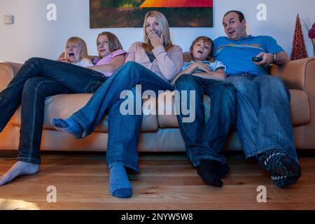 Family Life, TV Drama. Émotions mitigées sur le visage d'une jeune famille sur un canapé regardant la télévision ensemble à la maison. À partir d'une série d'images associées. Banque D'Images