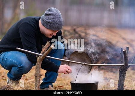 Un jeune homme prépare une soupe dans un chou-fleur suspendu au-dessus d'un feu, en l'remuant avec une grande cuillère. Banque D'Images