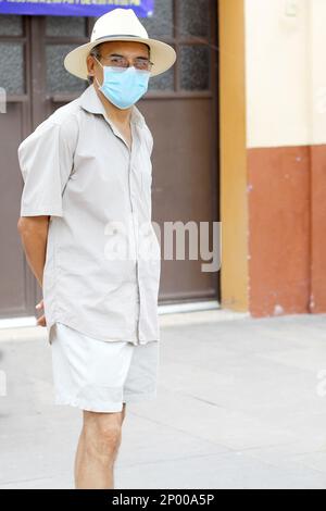 Latino adulte plus âgé avec masque de protection et chapeau marchant sur plaza, nouveau normal Covid-19 Banque D'Images