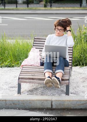 Elle est assise avec un ordinateur portable sur un banc de parc urbain. Freelance au travail. L'étudiant apprend à distance depuis l'extérieur. Style de vie moderne. Banque D'Images