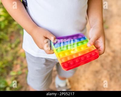 Petit garçon joue en caoutchouc carré jouet de violon Pop IT. Jouet antistress et tactile de couleur arc-en-ciel pour tous les âges. Loisirs en extérieur avec jouet pour moteur fin Banque D'Images