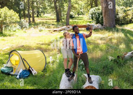 Couple heureux, divers en vacances de camping prenant selfie debout sur des rondins par tente dans la campagne Banque D'Images
