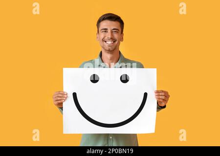 Jeune homme souriant tenant du papier avec une émoticône heureuse sur fond jaune Banque D'Images