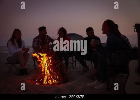 Groupe d'amis se rassemblant autour d'un feu de joie sur la plage le soir. Saison de camping Banque D'Images