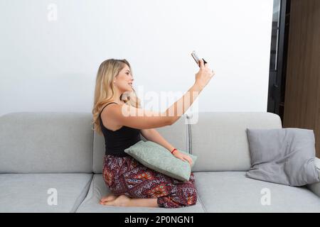 jolie femme blonde prenant des photos avec son smartphone sur le canapé de son salon Banque D'Images