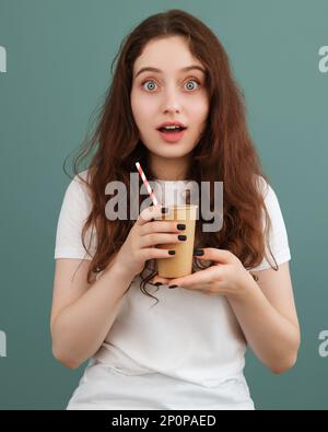 Une fille émervetée avec de grands yeux bleus tient une tasse en papier avec une paille rayée sur fond sarcelle. Copier l'espace sur le côté gauche. Banque D'Images