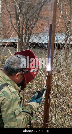 Un soudeur dans une veste de camouflage et un masque de protection rouge avec l'inscription en russe « masque de soudage F5 » travaille avec une machine à souder Banque D'Images