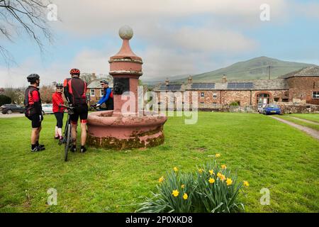 Cyclistes par la vieille fontaine d'eau sur le vert du village, avec Dufton Pike derrière, Dufton, Eden Valley, Cumbria. Fontaine d'eau construite par London Lead Co. Banque D'Images