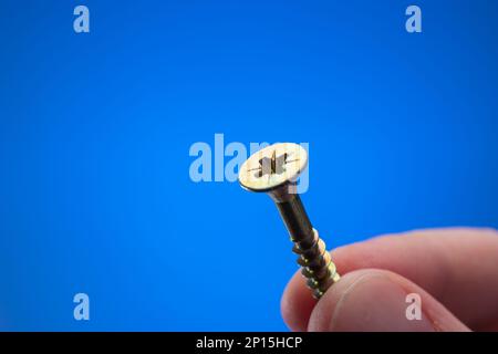 Petite vis filetée en métal maintenue entre les doigts par une main mâle caucasienne. Gros plan studio macro, isolé sur fond bleu Banque D'Images