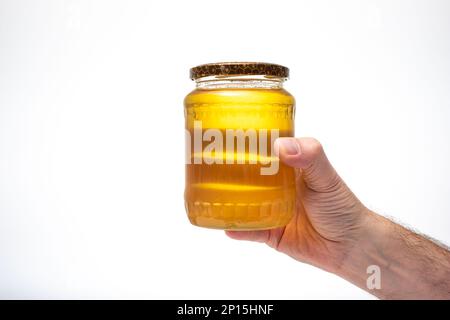 Miel d'abeille jaune d'or frais dans un pot en verre, tenu par la main mâle caucasienne. Gros plan en studio, isolé sur fond blanc. Banque D'Images