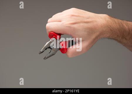 Main mâle caucasienne tenant une pince métallique avec poignées en plastique rouge isolées sur fond gris. Banque D'Images