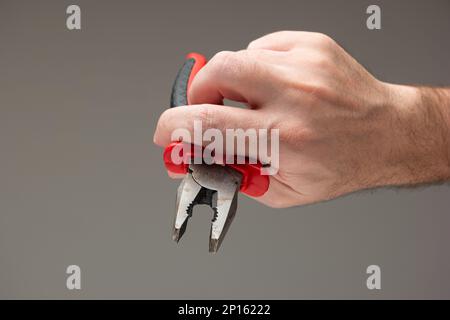 Main mâle caucasienne tenant une pince métallique avec poignées en plastique rouge isolées sur fond gris. Banque D'Images