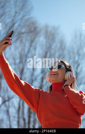 Jeune femme brune souriant avec un casque, des lunettes de soleil et un téléphone portable dans sa main, vêtue d'un chandail orange et d'un Jean Banque D'Images