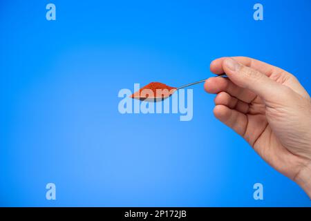 Poudre de paprika rouge hongrois dans une cuillère à thé métallique tenue à la main par un mâle caucasien isolé sur fond bleu prise en studio. Banque D'Images