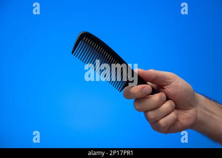 Main mâle caucasienne tenant un peigne à cheveux en plastique noir isolé sur fond bleu prise en studio. Banque D'Images