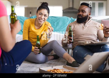 Des amis heureux et variés assis sur le sol, manger de la pizza et boire de la bière Banque D'Images