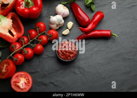 Délicieuse sauce adjika dans un bol et ingrédients sur une table noire, à plat Banque D'Images