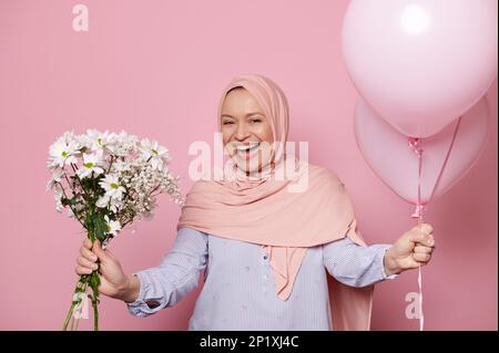 Une femme musulmane heureuse dans le hijab, rires regardant la caméra, tient un bouquet de fleurs blanches et ballons d'hélium, fond rose Banque D'Images