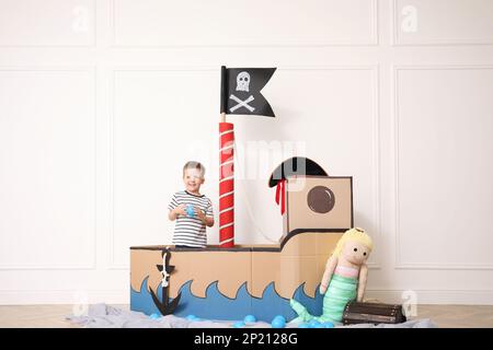 Mignon petit garçon jouant dans un bateau en carton pirate près du mur blanc à l'intérieur Banque D'Images