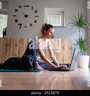 Jeune femme assise sur le sol d'un salon utilisant ou travaillant sur un ordinateur portable. Son chien d'animal de compagnie Labrador retriever noir allongé à côté d'elle sur un tapis. Banque D'Images