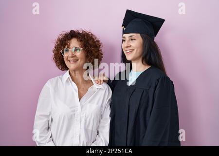 Mère et fille hispaniques portant une casquette de remise des diplômes et une robe de cérémonie en regardant de côté avec le sourire sur le visage, expression naturelle. rire confiant. Banque D'Images