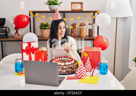 Femme hispanique célébrant l'anniversaire avec un grand gâteau au chocolat faisant appel vidéo pointant le pouce vers le côté souriant heureux avec la bouche ouverte Banque D'Images