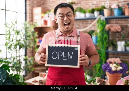 Jeune homme chinois travaillant chez un fleuriste tenant un panneau ouvert célébrant fou et stupéfié pour le succès avec les yeux ouverts hurlant excitant. Banque D'Images