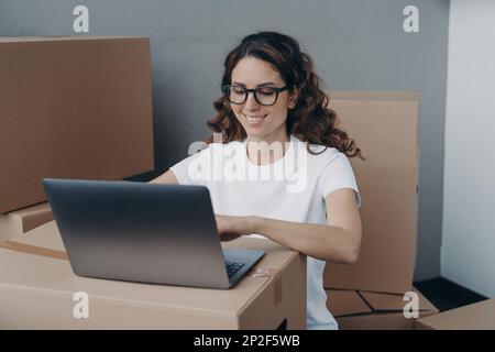 Femme hispanique souriante portant des lunettes travaillant sur un ordinateur portable entouré de boîtes en carton, préparant les commandes des clients, colis de la boutique en ligne pour sen Banque D'Images