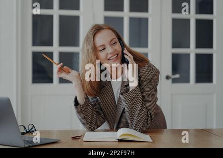 Une jeune femme d'affaires souriante parle au téléphone pour écrire des notes, assise au bureau. Femme secrétaire professionnelle assistante personnelle tenant un smartphone Banque D'Images