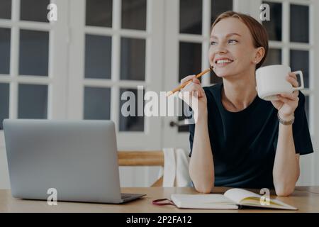 Femme pensive souriante tenant un crayon et une liste d'écriture de tasse dans un carnet, assis au bureau, regardant de côté, rêvant, pensant. Des notes de femme rêveuses Banque D'Images