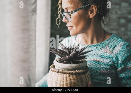 Vue latérale d'une femme pensive tenant l'usine à l'intérieur de la maison et regardant à l'extérieur de la fenêtre seule. Femmes portant des lunettes et un intérieur protecteur Banque D'Images