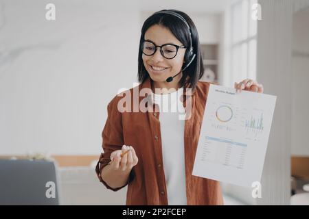 Une femme souriante dans un micro-casque/une oreillette affiche un document avec des statistiques lors d'une conférence en ligne sur un ordinateur portable. Femme d'affaires amicale dans le casque ayant la vidéo Banque D'Images