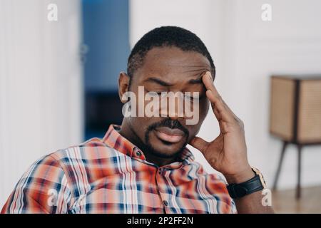 L'homme afro a eu mal de tête et se sent endormi. Visage de l'enseignant en ligne fatigué qui a fermé les yeux et touche la tête. Travaillez à domicile ou à distance le soir Banque D'Images