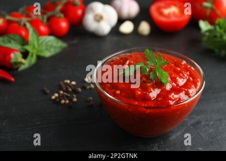 Délicieuse sauce adjika avec persil dans un bol en verre et ingrédients sur une table noire, en gros plan Banque D'Images