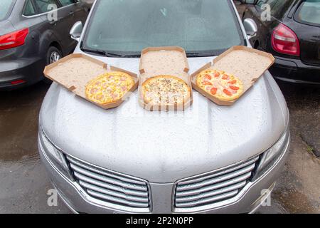 Trois pizzas dans des boîtes à emporter ouvertes en carton sont situées sur le capot d'une voiture dans le parking. Un ensemble de trois pizzas pour le déjeuner par un groupe de travée Banque D'Images