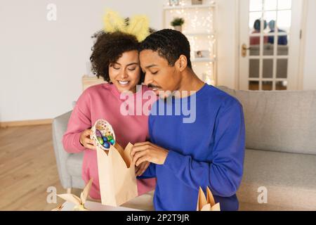 Joyeux jeune Noir couple célébrant Pâques à la maison, faisant des emballages cadeaux de vacances Banque D'Images