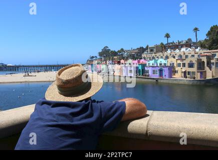 Un homme âgé portant un chapeau de paille se détend sur le balcon d'un restaurant donnant sur les cottages colorés de l'hôtel à Capitola, CA. Banque D'Images