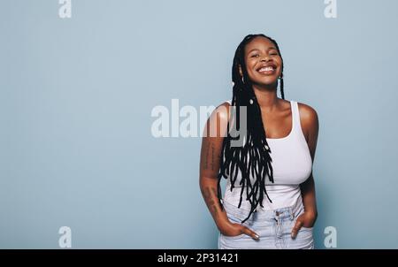 Jeune femme souriant à la caméra tout en se tenant debout sur un fond bleu. Jeune femme noire se sentant confiante dans son style. Banque D'Images