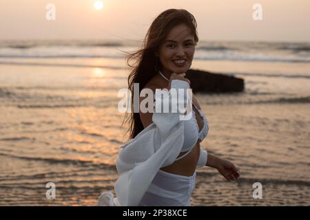 Portrait de jeune belle brunette souriante avec des cheveux longs ample marchant le long de la mer au coucher du soleil. Vacances en mer. Banque D'Images