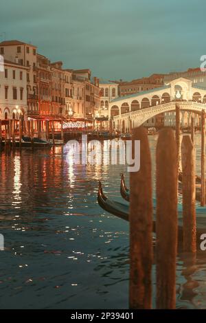 Des poteaux d'amarrage et une gondole encadrent le pont du Rialto illuminé enjambant le Grand Canal au crépuscule à Venise, Italie, rendu dans des tons vintage chaleureux. Banque D'Images