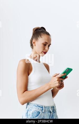 Une jeune femme à la belle peau bronzée regarde l'écran du téléphone dans ses mains en portant un T-shirt blanc et un Jean bleu contre un mur blanc Banque D'Images