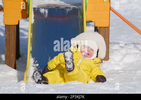 Un tout-petit garçon joue sur un terrain de jeu en hiver. Un enfant dans une combinaison jaune sur une glissade d'enfant dans la neige. Enfant un an e Banque D'Images