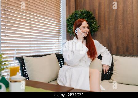 Portrait d'une jeune femme souriante portant un peignoir blanc, assise sur un canapé confortable près de la fenêtre et parlant sur un smartphone, en appréciant la conversation Banque D'Images