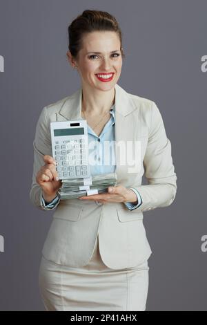 heureuse femme d'affaires moderne de 40 ans dans un costume d'affaires léger avec calculatrice et dollars packs d'argent sur fond gris. Banque D'Images