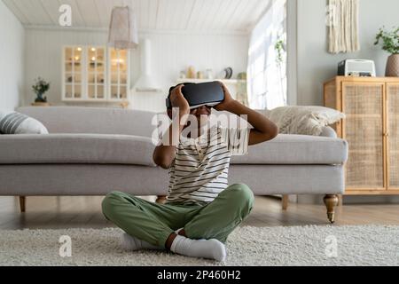 Curieux Afro-américain garçon avec des lunettes VR assis sur le sol simulation d'étude monde virtuel à la maison Banque D'Images