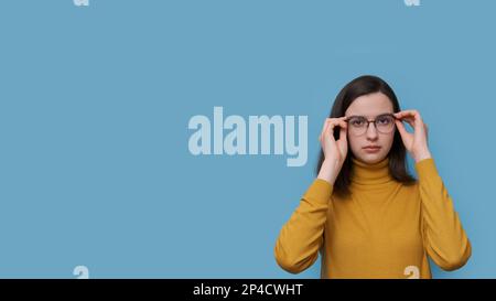 Une jeune fille étudiante souriante qui prend ou met des lunettes sur un fond bleu. La fille voit mieux que d'essayer de nouvelles lunettes prescrites. vérification du soupir Banque D'Images