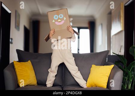 Portrait de style de vie d'un enfant jouant avec une boîte en carton sur le canapé dans la salle de séjour. Mignon enfant portant un costume robot à la maison Banque D'Images