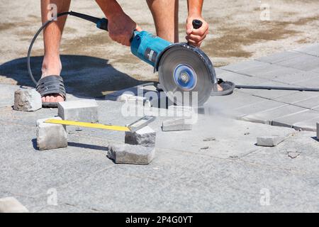 Les mains d'un travailleur sur la rue ont coupé des dalles de pavage avec une meuleuse pour le pavage ultérieur du trottoir lors d'une journée ensoleillée d'été. Gros plan, espace de copie. Banque D'Images